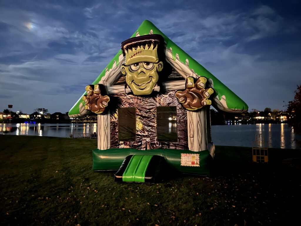 Inflatable Frankenstein-themed bounce house with a green roof, large 3D Frankenstein face, and outstretched hands, set up at night near a lake.
