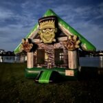 Inflatable Frankenstein-themed bounce house with a green roof, large 3D Frankenstein face, and outstretched hands, set up at night near a lake.